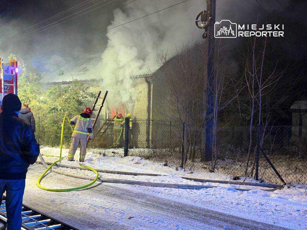 Strażacy gaszą pożar budynku, otoczeni dymem w zimowym otoczeniu, obok widoczni zaniepokojeni obserwatorzy.
