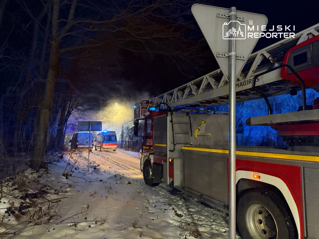 W nocy samochód strażacki z drabiną oraz karetka na zaśnieżonej drodze, w tle dym i niebieskie światła.