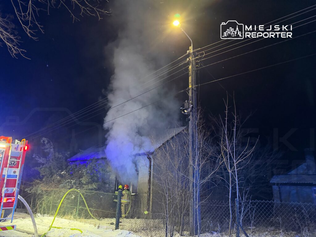 Strażacy gaszą pożar w drewnianym budynku, z którego wydobywa się dym, w otoczeniu zimowej scenerii.