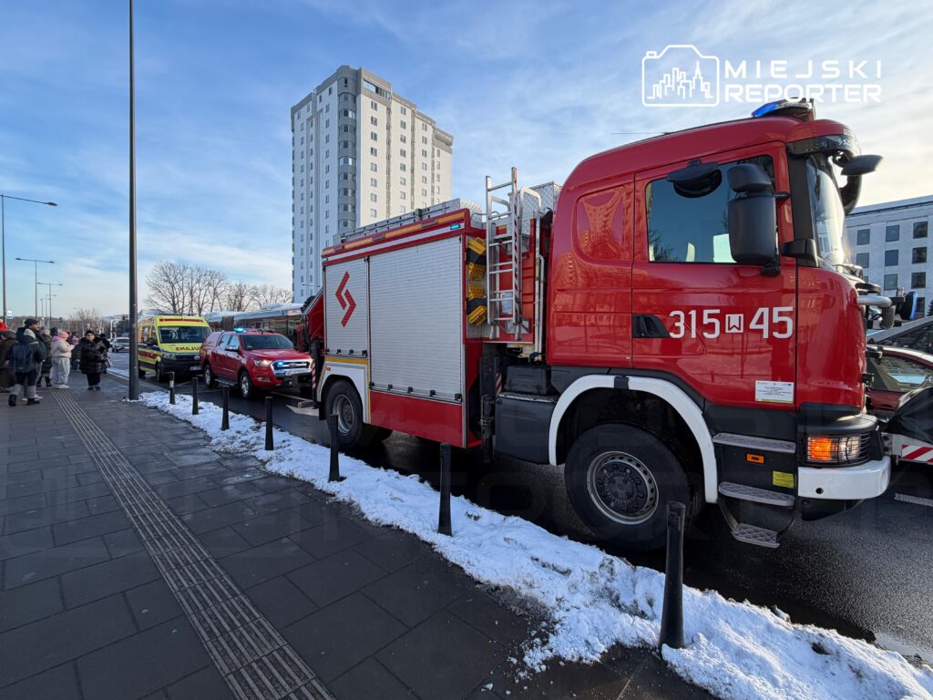 Czerwony wóz strażacki z otwartą klapą stoi na ulicy, obok niego zaparkowany czerwony samochód osobowy, a w tle grupa ludzi obserwujących sytuację.