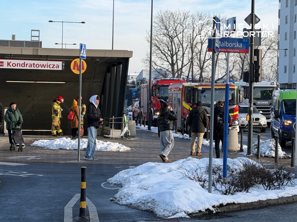 Grupa strażaków w mundurach zjeżdża się do stacji metra, obok ludzi czekających na przystanku, w zimowej scenerii.