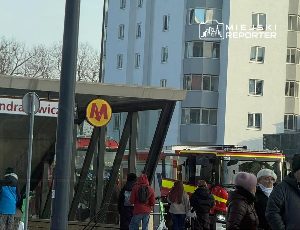 Ludzie czekają przy stacji metra z widocznym znakiem "M", obok podjeżdża wóz strażacki.