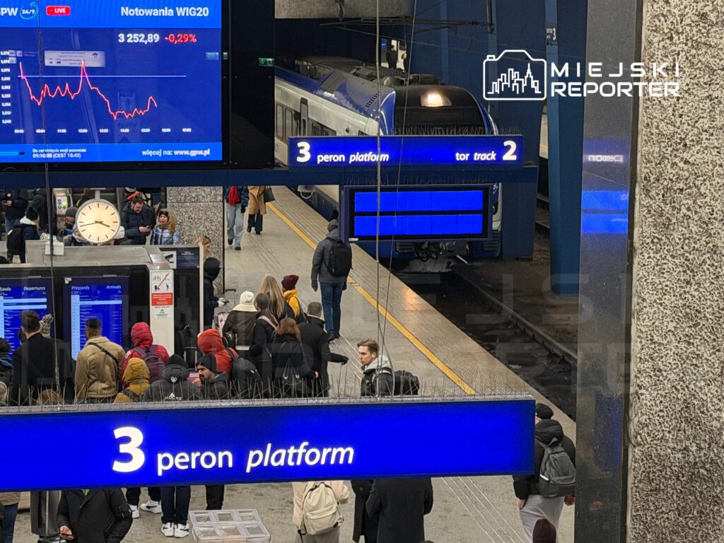 Tłum ludzi czeka na pociąg na trzecim peronie stacji kolejowej, w tle widoczny pociąg i zegar.