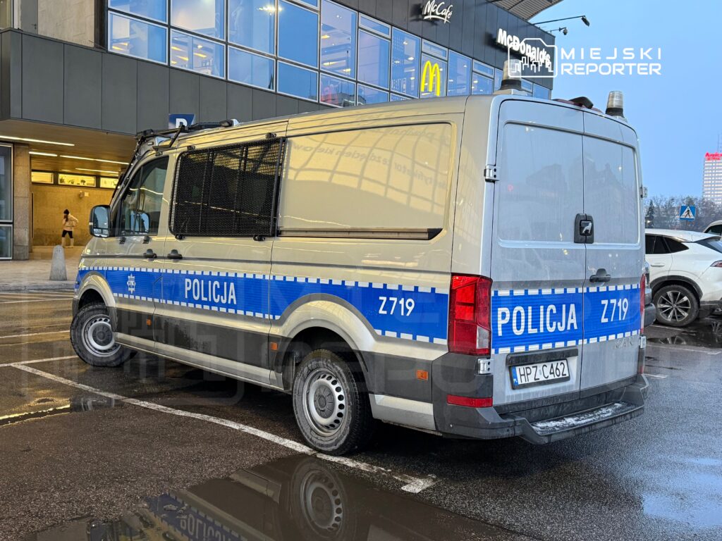 Policyjny van zaparkowany na ulicy przed nowoczesnym budynkiem handlowym w deszczowy dzień.