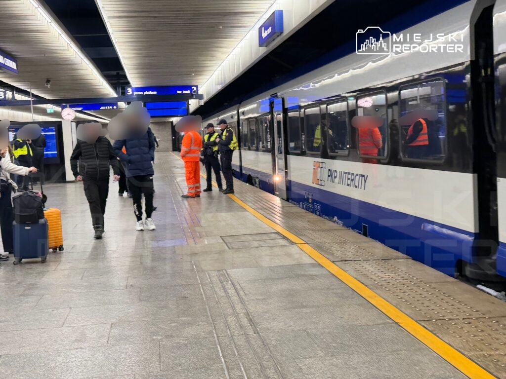 Pasażerowie z bagażami czekają na peronie, obok stoją funkcjonariusze w uniformach oraz pracownicy w pomarańczowych kamizelkach.