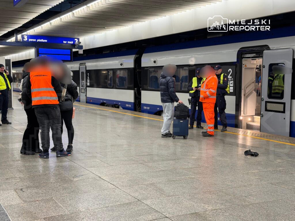 Grupa osób w odblaskowych kamizelkach rozmawia z mężczyzną z walizką na peronie stacji kolejowej.