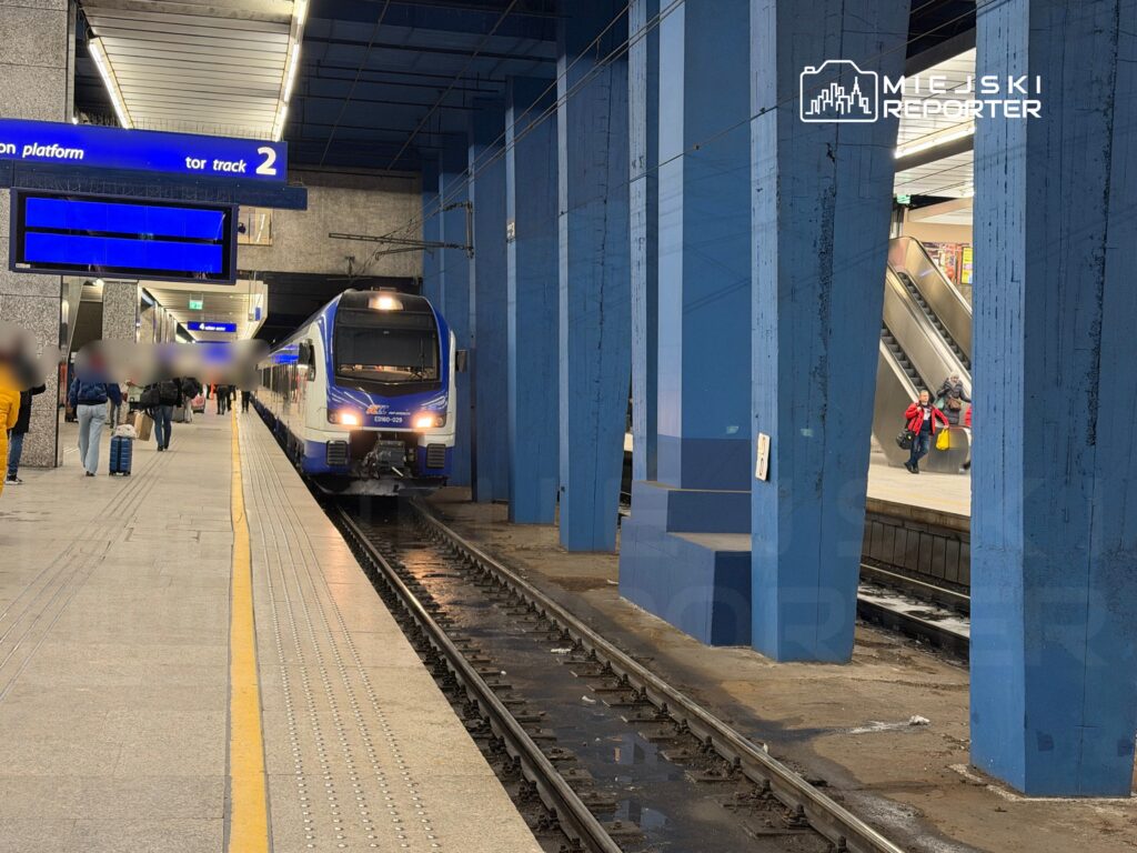 Pociąg osobowy zatrzymany na peronie z oznaczeniem "tor track 2", obok pasażerowie czekają na przyjazd.