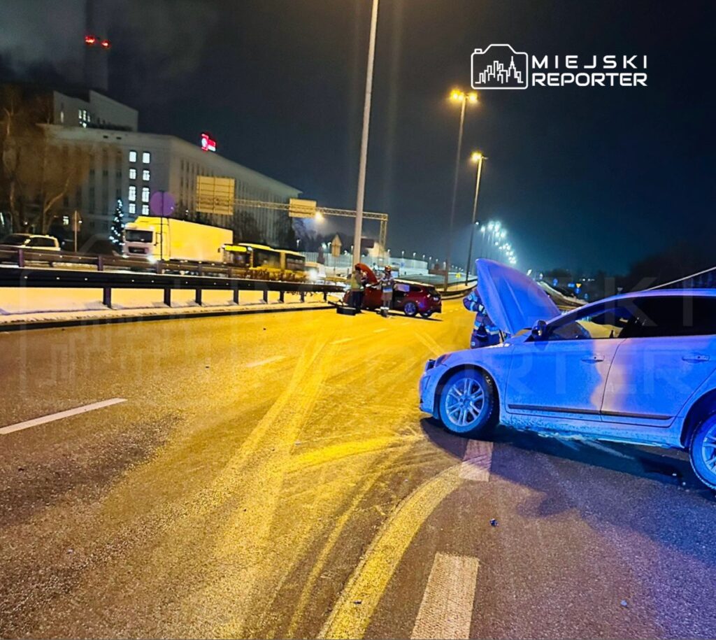 Dwa uszkodzone samochody na jezdni, jeden z otwartą maską, w nocy na drodze z widocznymi śladami poślizgu.