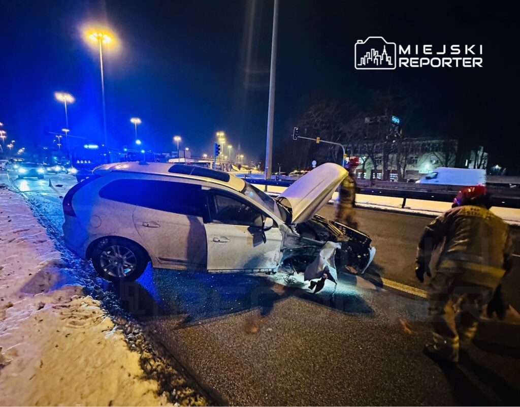 Zniszczony samochód osobowy na jezdni, przy którym interweniują strażacy, w nocy, w oświetlonym mieście.