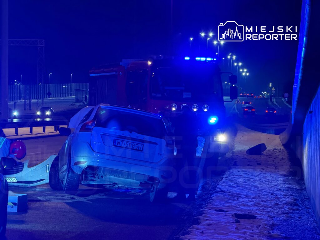 W nocy na drodze stoi uszkodzony samochód osobowy, a obok niego pracują strażacy z wozem ratunkowym.