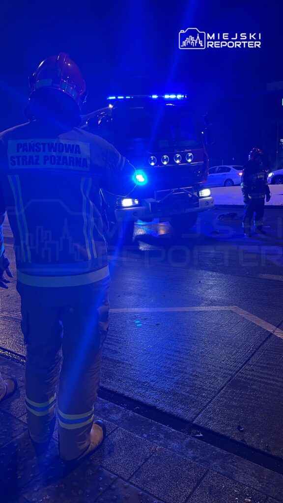 Strażak w mundurze z odblaskowymi elementami monitoruje sytuację przy włączonym wozie strażackim z niebieskimi światłami.