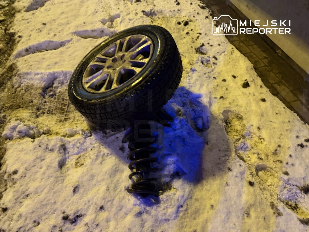 Odpadające koło z oponą leży na zaśnieżonym chodniku obok metalowego sprężynowego zawieszenia.