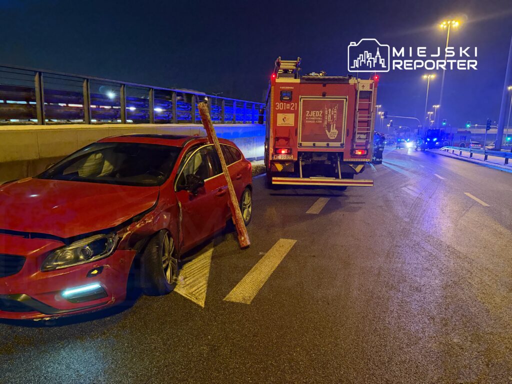 Czerwony samochód osobowy z uszkodzeniami stoi na drodze, obok niego wóz strażacki podczas akcji ratunkowej w nocy.