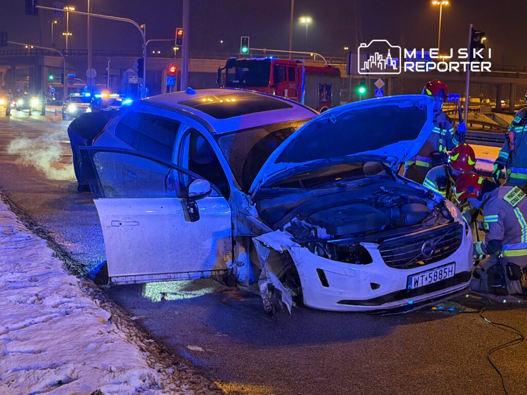 Uszkodzony samochód osobowy z otwartą maską na drodze, obok pracujący strażacy w hełmach i mundurach.
