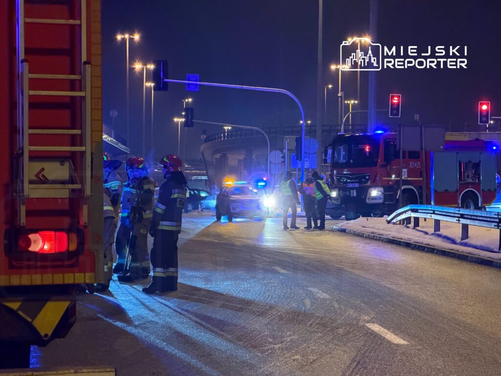 Grupa strażaków w hełmach omawia sytuację na oświetlonej nocą drodze, obok wozu strażackiego i radiowozu.