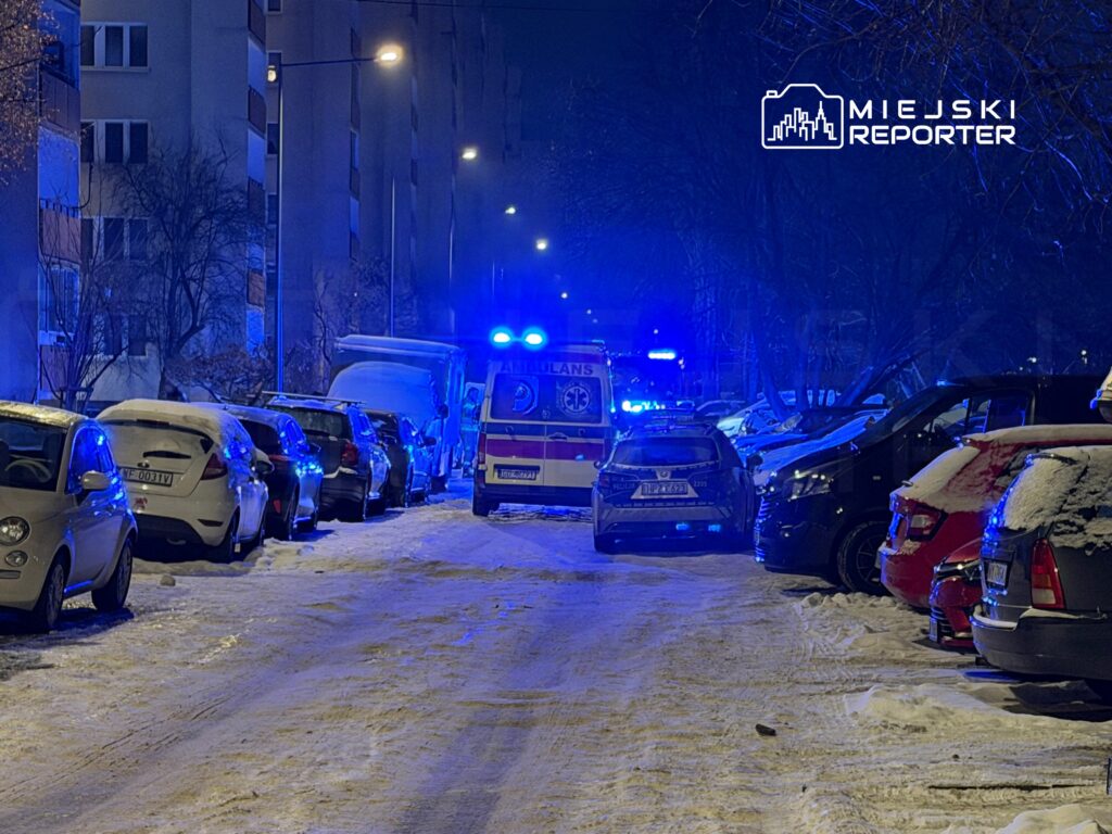 Nocna ulica pokryta śniegiem, z radiowozem i karetką na końcu, oświetlona niebieskimi światłami.