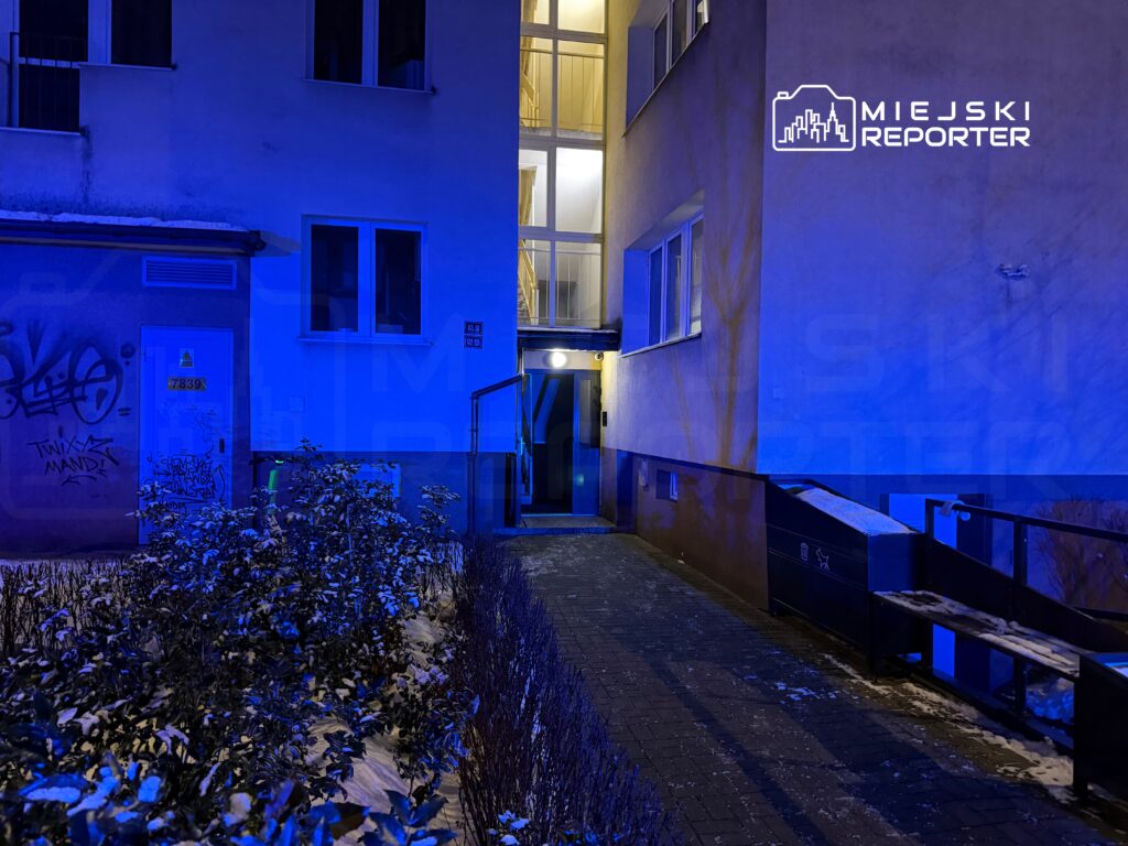 Nocna sceneria przy budynku mieszkalnym o niebieskim oświetleniu, z graffiti na ścianie i ścieżką prowadzącą do wejścia.