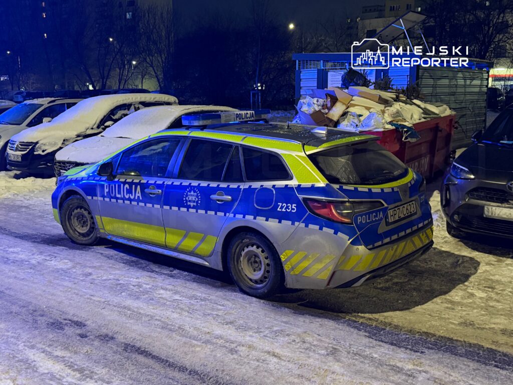 Policyjny samochód zaparkowany na ośnieżonym parkingu, obok innych pojazdów, w nocy.