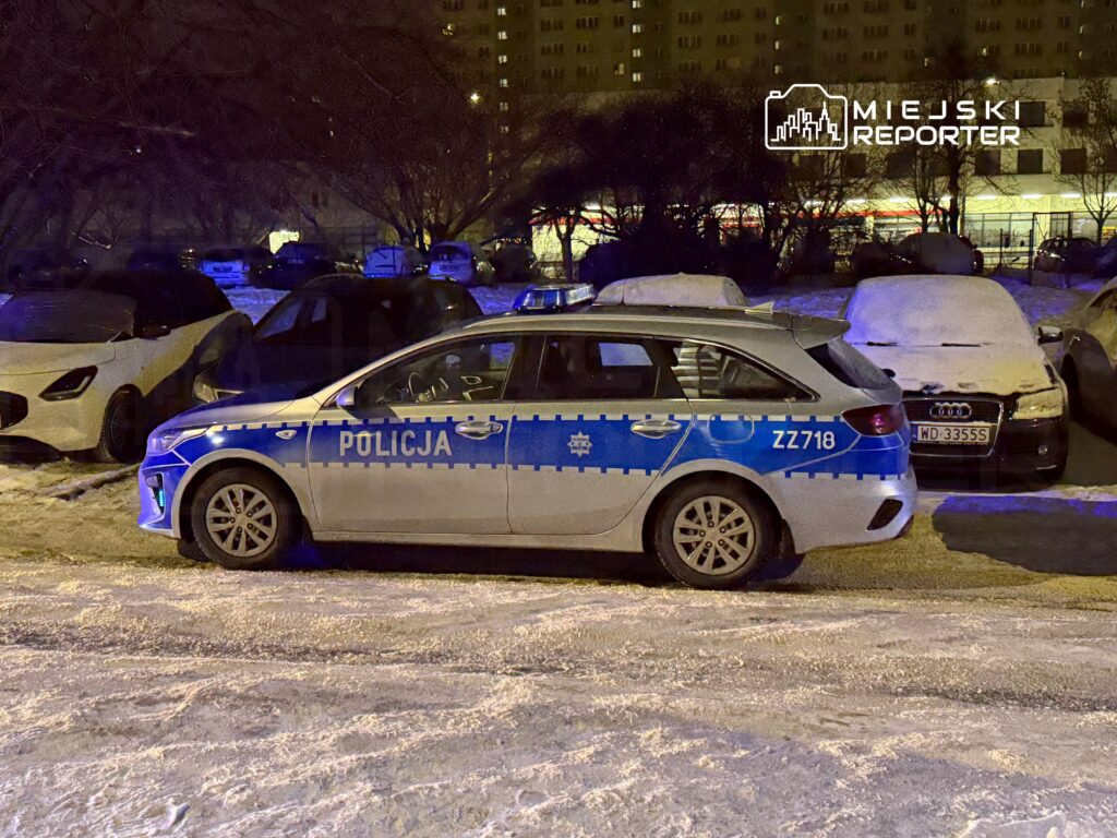 Radiowóz policyjny z oznaczeniem "POLICJA" zaparkowany na zaśnieżonym parkingu nocą, w tle inne samochody.