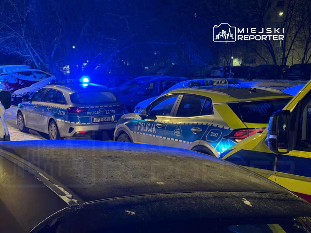 Policyjne samochody zaparkowane w oświetlonym niebieskim świetle, otoczone przez inne pojazdy w zimowej scenerii.