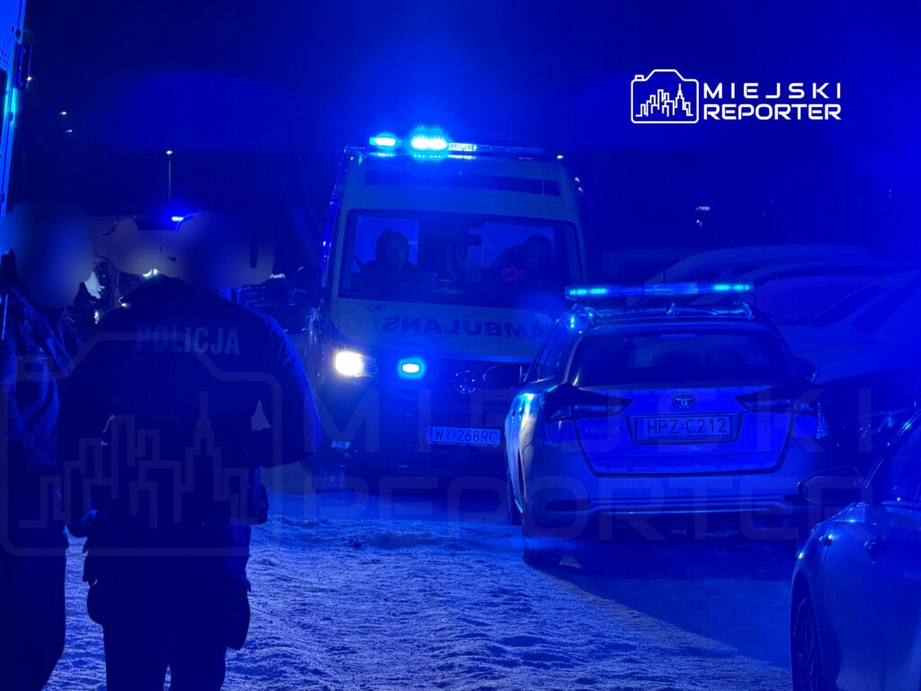 Policjant w kurtce z napisem "POLICJA" rozmawia z osobami w zaśnieżonym terenie, w tle widać radiowóz i karetkę.