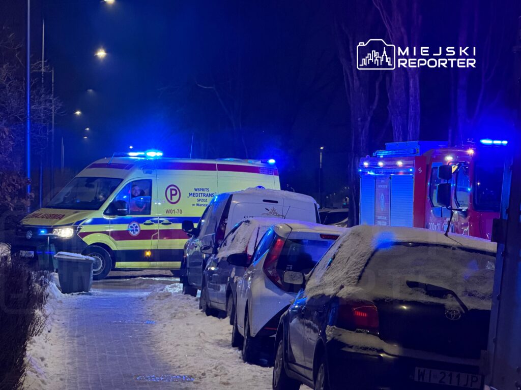 Karetka pogotowia z włączonymi sygnałami świetlnymi stoi na ośnieżonej ulicy, obok zaparkowanych samochodów.