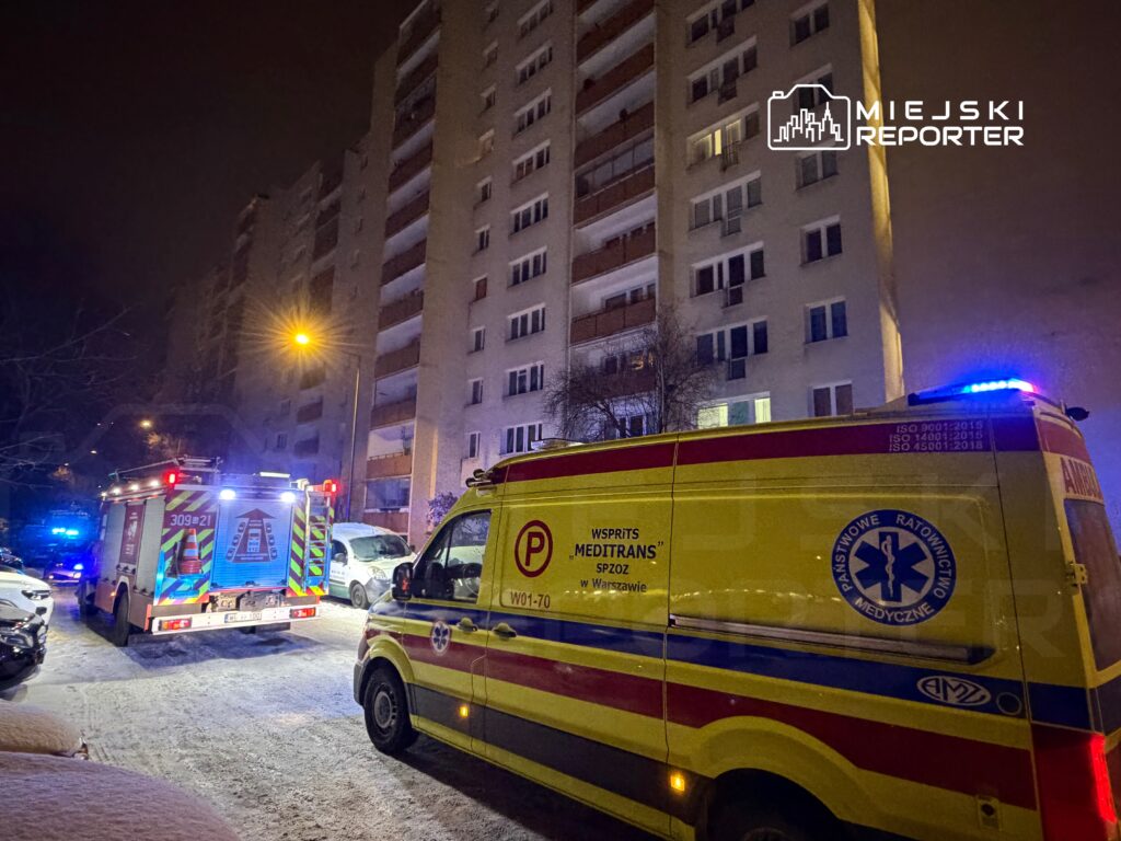Karetka pogotowia oraz wóz strażacki stoją na ośnieżonej ulicy przed wysokim budynkiem mieszkalnym w nocy.