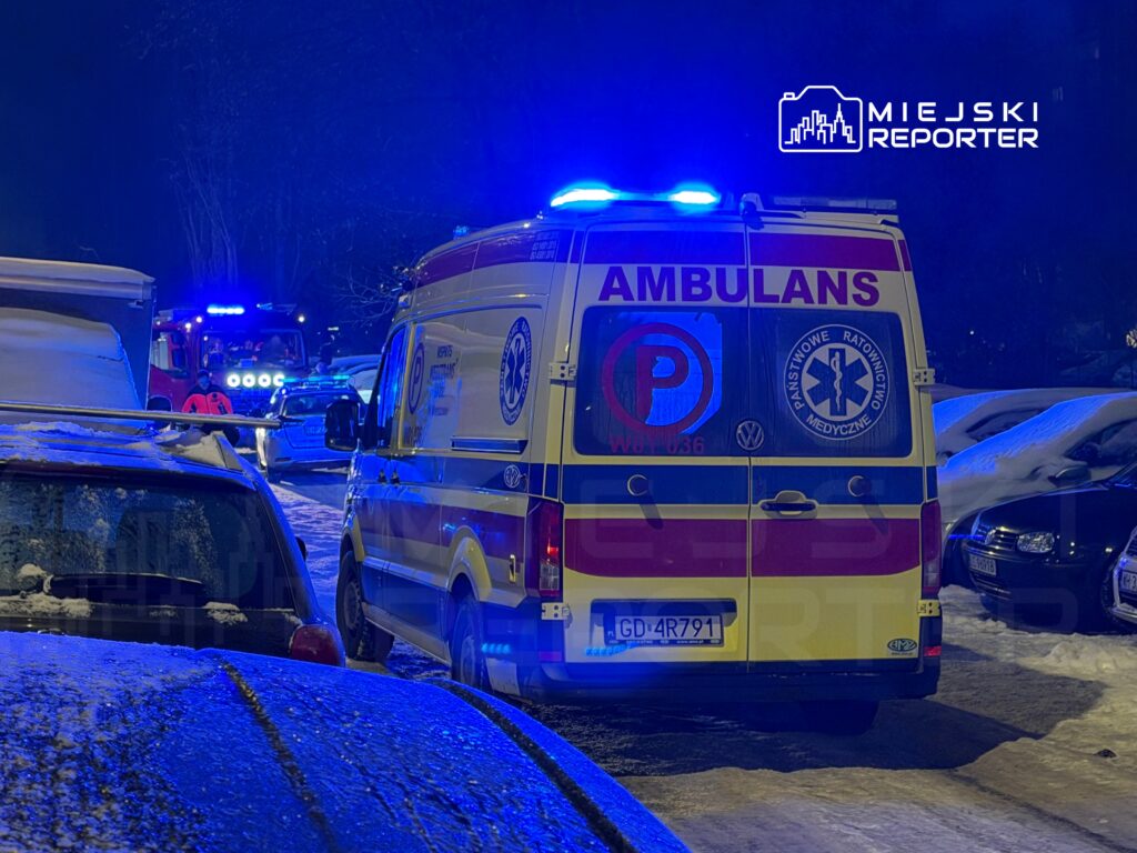 Karetka pogotowia z włączonymi sygnalizatorami świetlnymi stoi na zaśnieżonym parkingu nocą, w otoczeniu innych pojazdów.