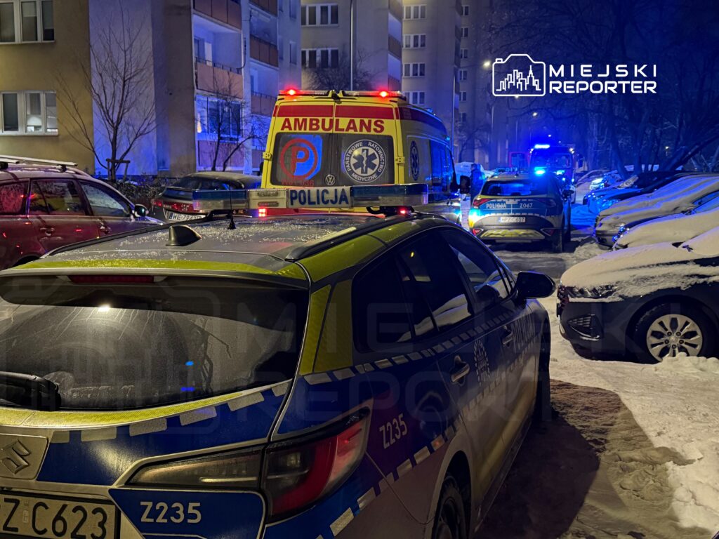 Karetkę pogotowia i radiowozy policji zaparkowane w ośnieżonym osiedlu mieszkaniowym w nocy.