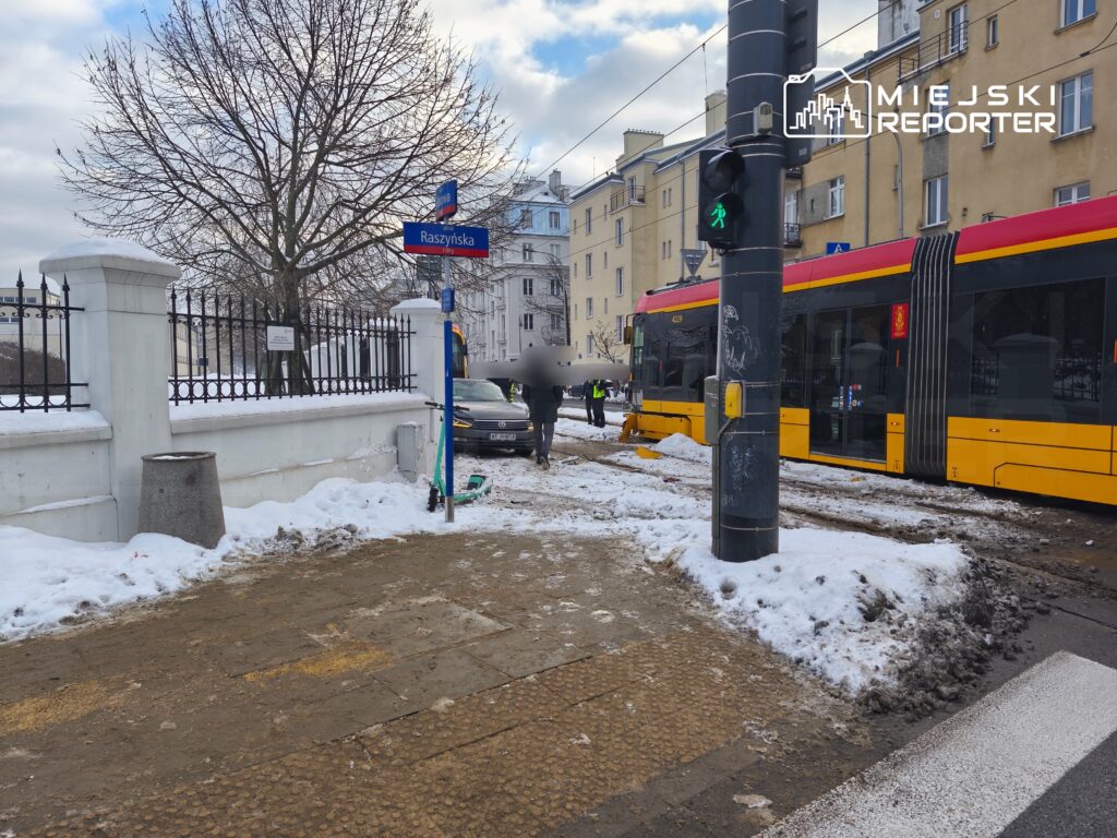 Ruchliwa ulica z tramwajem zatrzymującym się na przystanku, w tle widoczny znak uliczny "Raszynska".