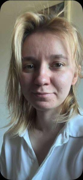 Kobieta z długimi, blond włosami, uśmiechająca się w jasnej bluzie, siedzi w domowym otoczeniu.