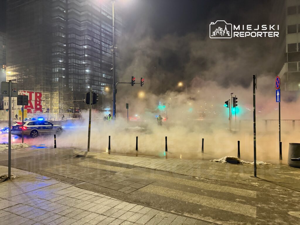 Policyjny radiowóz z niebieskimi światłami stoi na ulicy, w tle widoczna mgła oraz nowoczesny budynek.