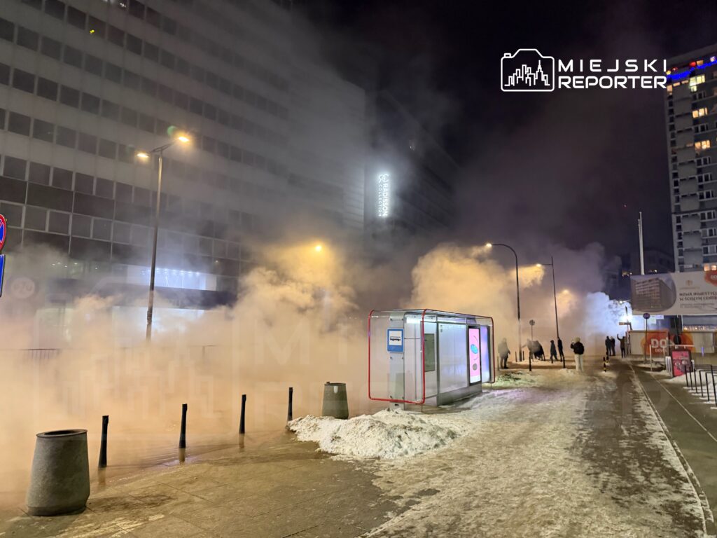 Gęsta mgła unosząca się nad chodnikiem w miejskim otoczeniu, z przystankiem autobusowym w tle.