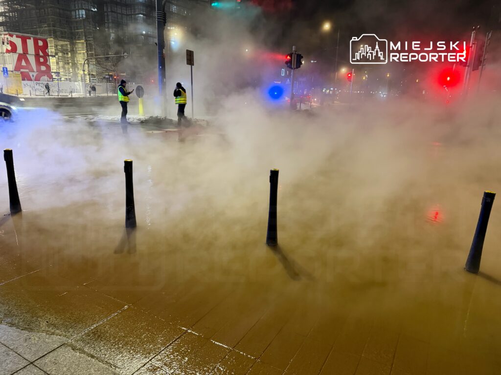Dwaj mężczyźni w odblaskowych kamizelkach stoją na mokrej ulicy, otoczeni gęstą mgłą.