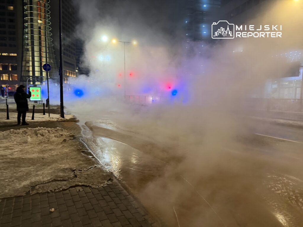 Mężczyzna w czarnej kurtce stoi na chodniku, otoczony gęstą mgłą, obok świecących sygnalizatorów i budynków.