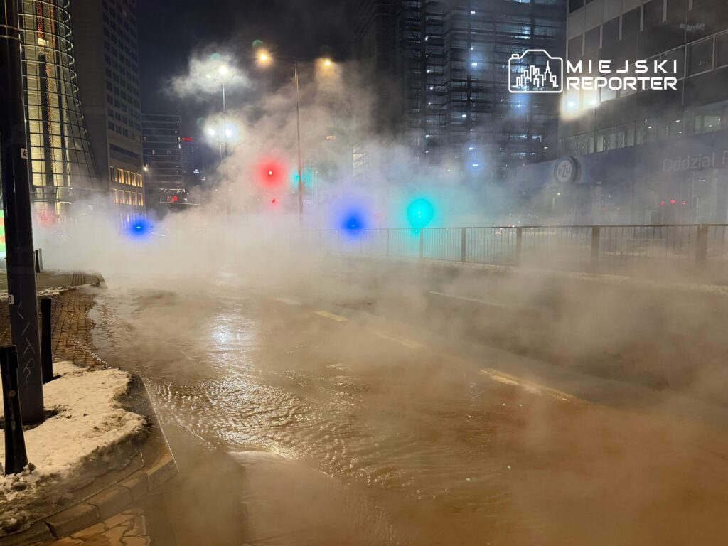 Gęsta mgła unosząca się nad zalaną ulicą, w tle świecące sygnalizatory świetlne i nowoczesne budynki.