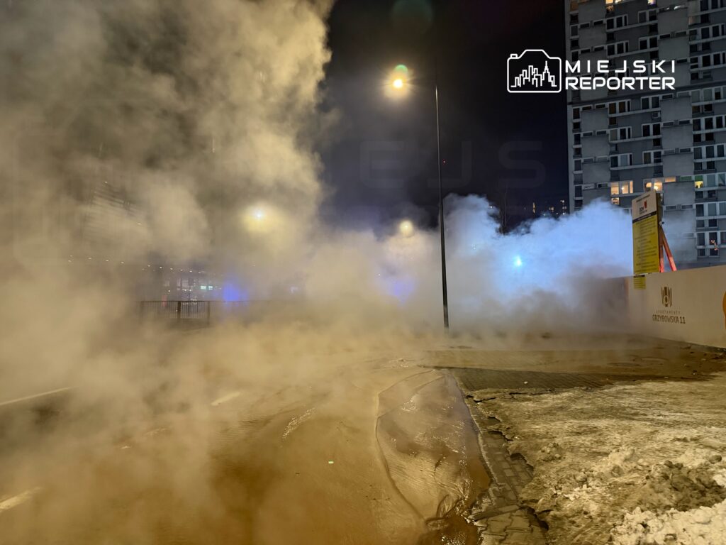 Gęsta mgła unosząca się na ulicy w nocy, z widocznymi latarniami i fragmentem budynku w tle.