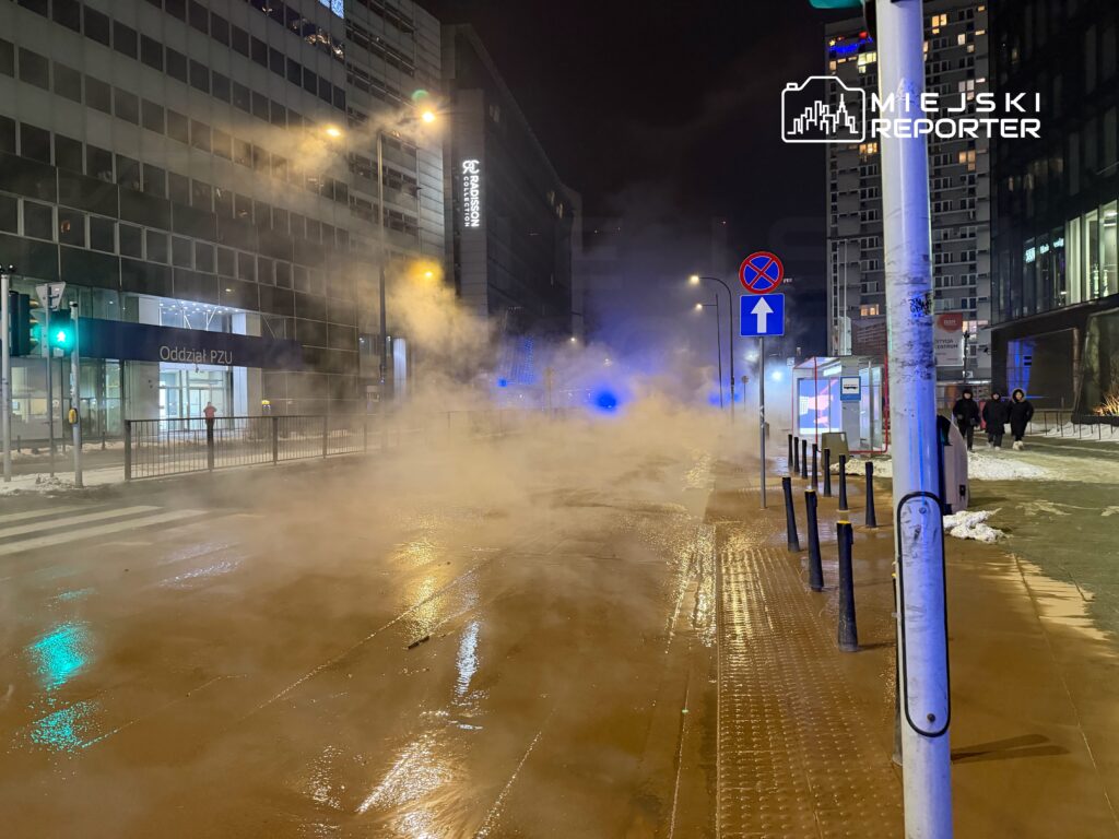 Ulica w nocy z widoczną mgłą pary, oświetlona zielonym światłem sygnalizatora, obok biurowca PZU.