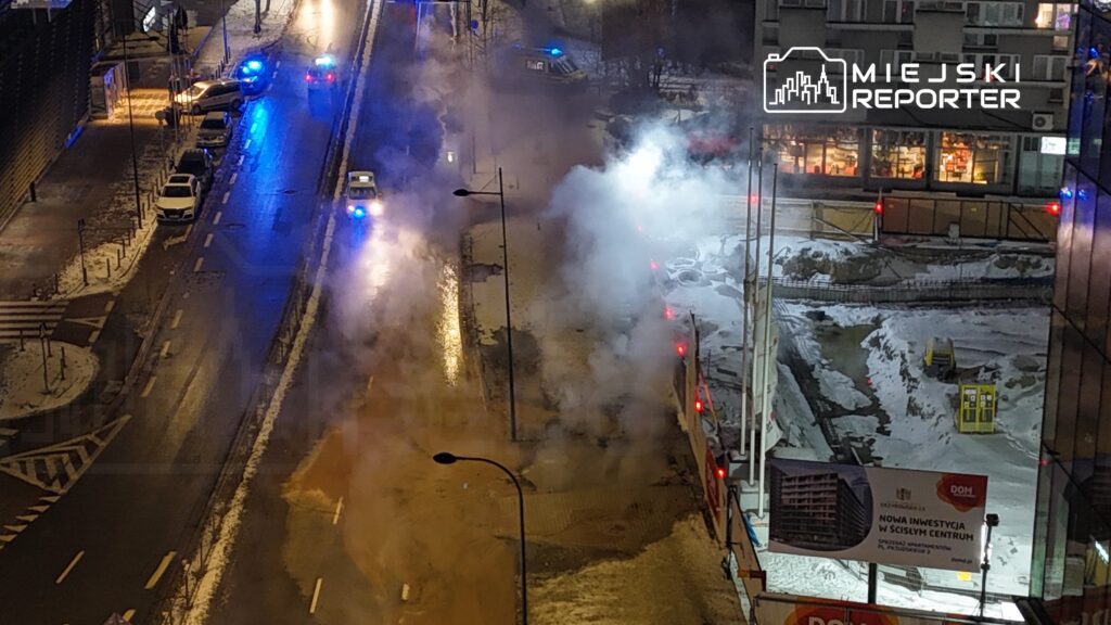 Na ulicy widać unoszący się dym oraz policyjne radiowozy z włączonymi sygnałami świetlnymi. W tle znajduje się plac budowy z widocznymi maszynami.