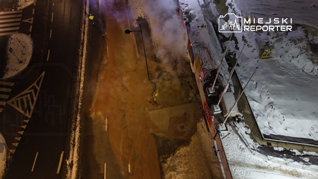 Para pracowników w odblaskowych kamizelkach ocenia wyciek wody na zaśnieżonej ulicy, obok widoczna para.