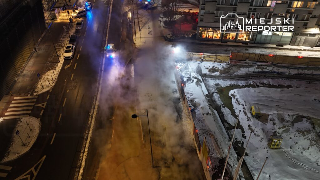 Na zaśnieżonej ulicy unosi się para, a w tle widać niebieskie światła radiowozów i budowę z ogrodzeniem.