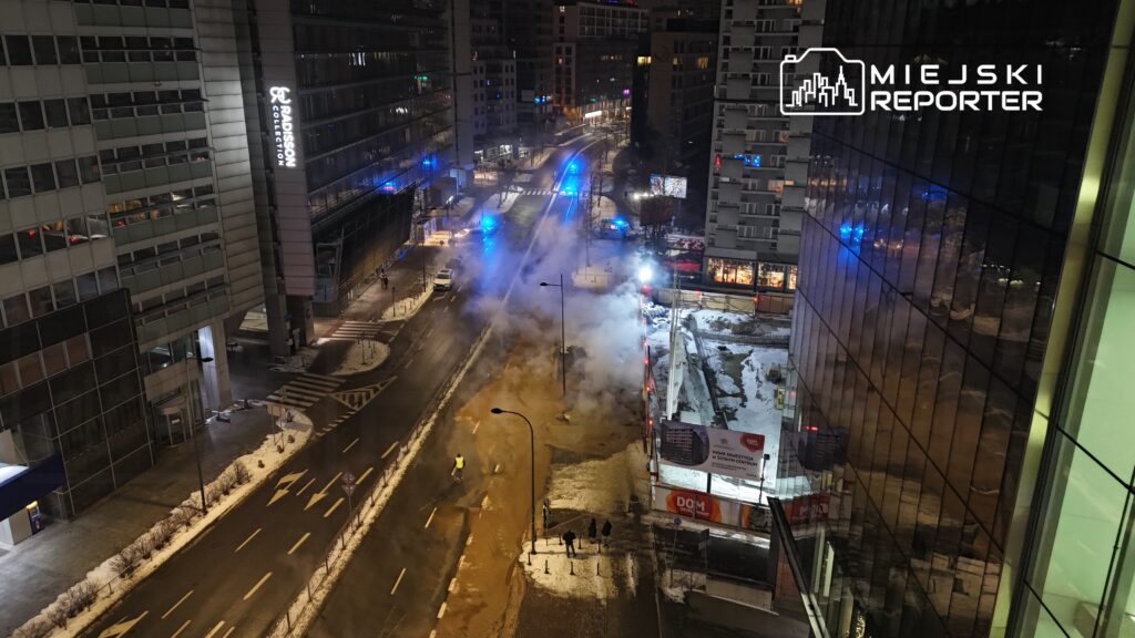 Na ulicy w nocy unosi się para z miejsca, gdzie występuje wyciek wody, a w tle widoczne są niebieskie światła policyjne.