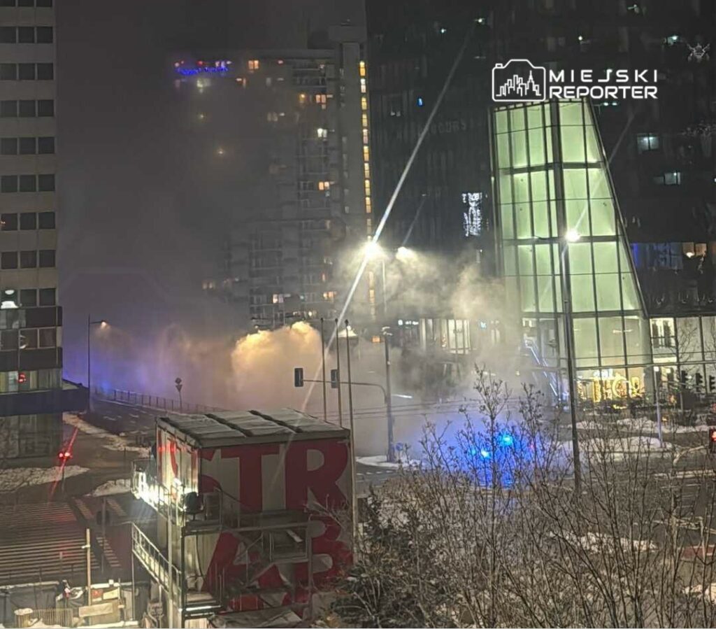 Gęsta mgła unosząca się w miejskim krajobrazie, z niebieskimi światłami radiowozu w tle.