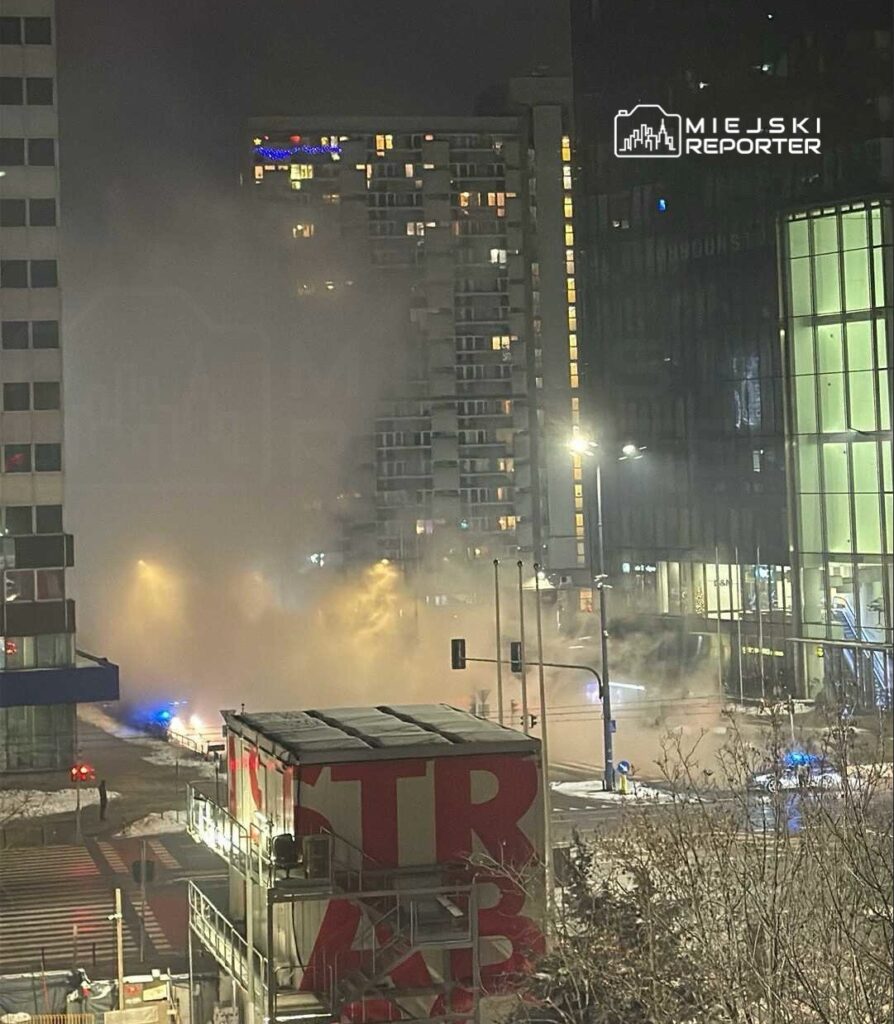 Mglista noc w mieście, z widocznymi światłami policyjnymi i parą unoszącą się nad ulicą. W tle nowoczesne budynki.