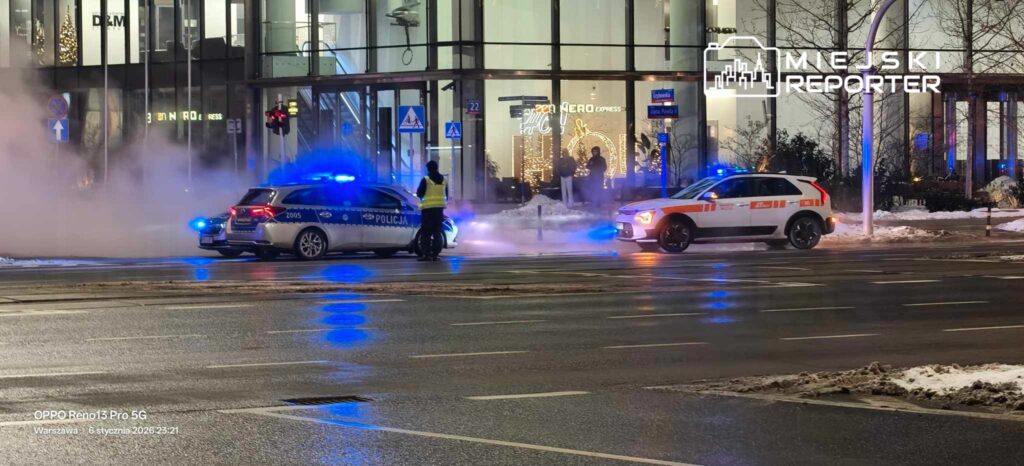 Policyjny samochód z włączonymi sygnałami świetlnymi zatrzymany na skrzyżowaniu, obok drugiego pojazdu w tle.