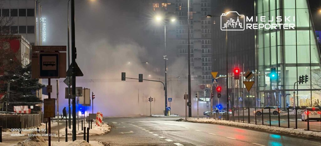 Zamglona ulica z widocznymi sygnalizatorami świetlnymi i policyjnym radiowozem w tle, w otoczeniu nowoczesnych budynków.