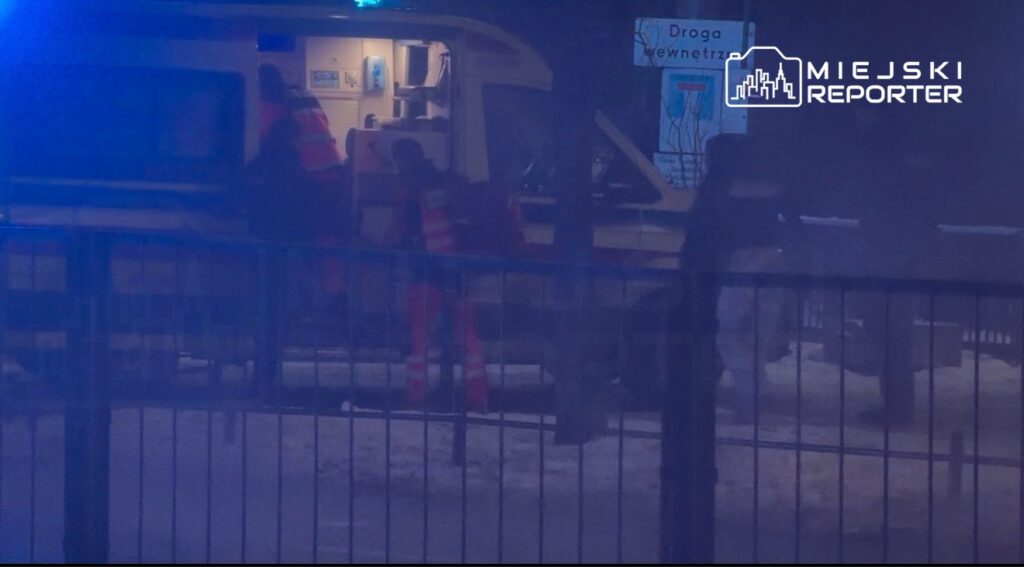 Ratownicy medyczni przenoszą osobę do karetki w zaśnieżonym otoczeniu, przy niebieskich światłach alarmowych.