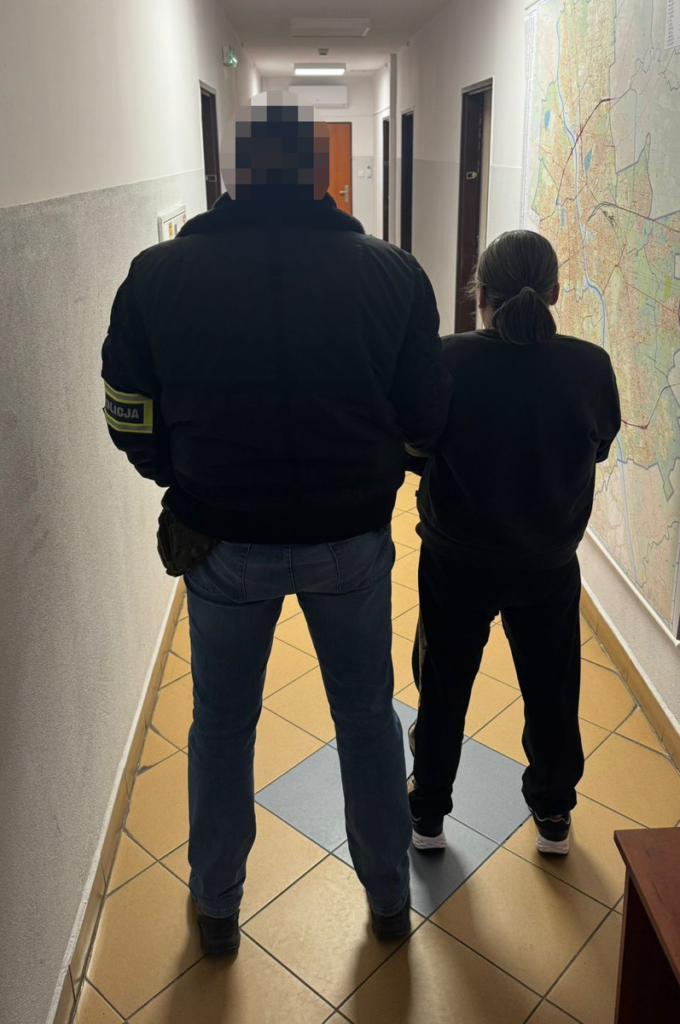 Funkcjonariusz policji prowadzi osobę w czarnym dresie korytarzem w budynku administracyjnym. Na ścianie mapa.