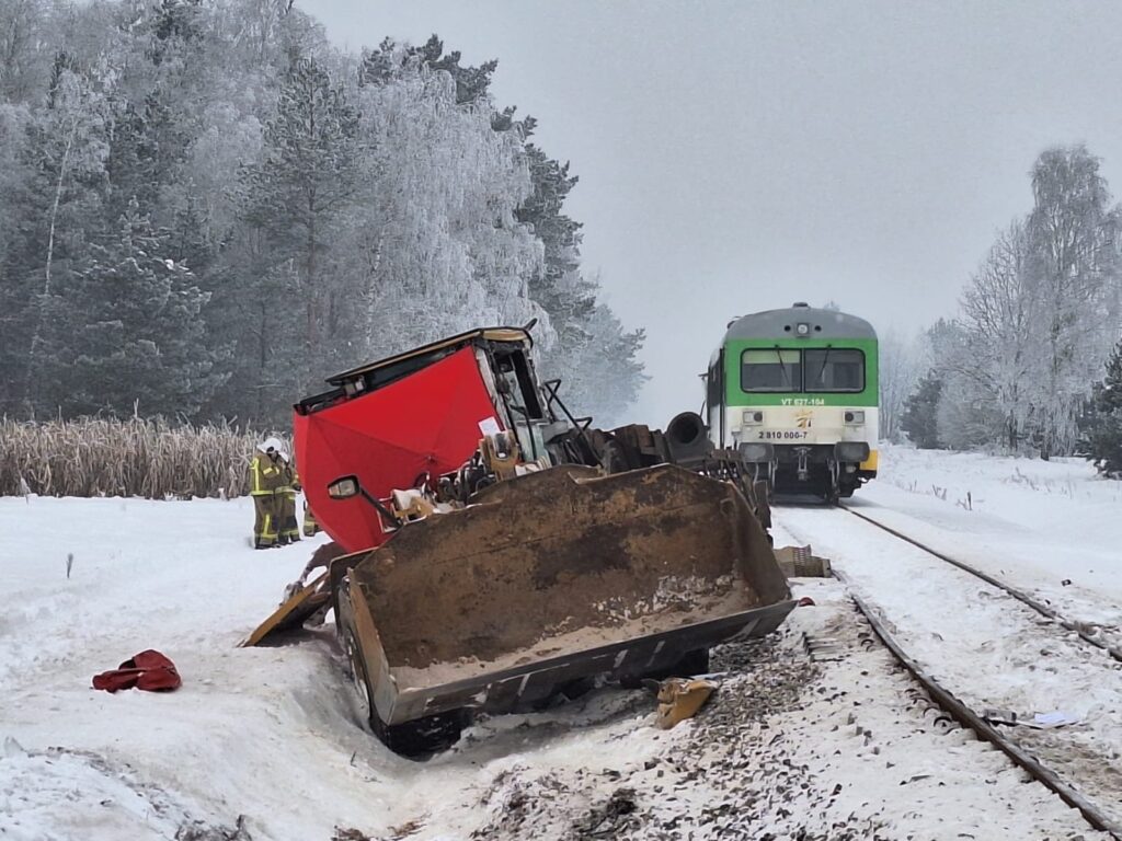 Wielka maszyna budowlana z czerwoną kabiną stoi w śniegu obok torów, a ratownicy rozmawiają w tle.