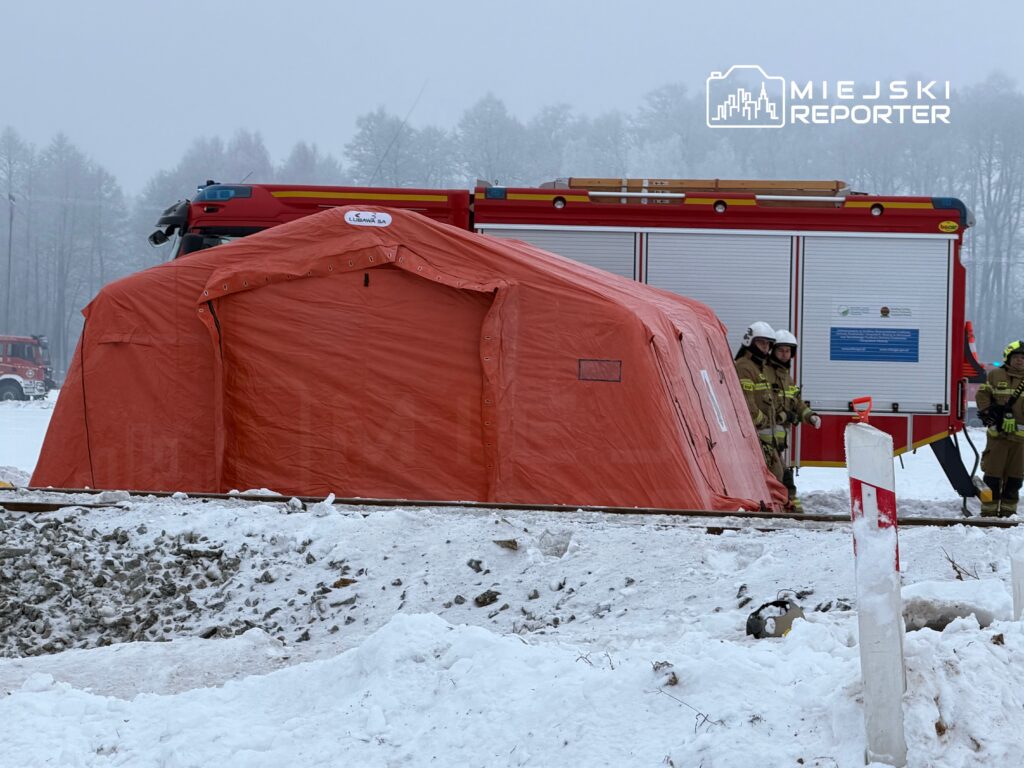 Strażacy w hełmach stoją obok pomarańczowego namiotu ratunkowego na zaśnieżonym terenie, w tle wóz strażacki.